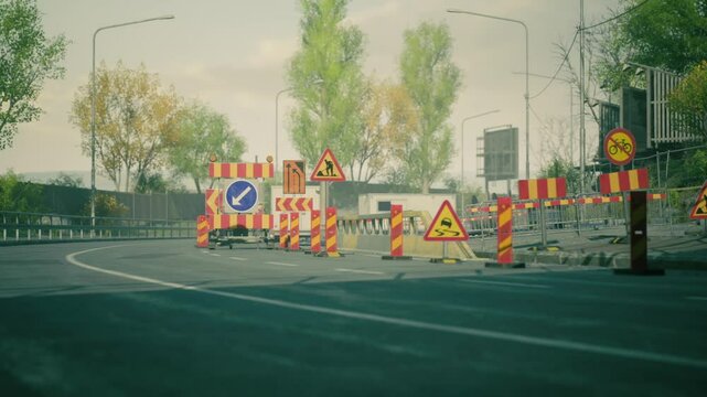 Workers are setting up detour signs and barriers for an ongoing road construction project in an urban area. The trees are changing colors, indicating autumn.