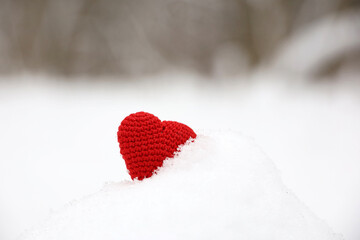 Fototapeta premium Red knitted heart in the snow on blurred forest background. Symbol of romantic love, Valentine's card