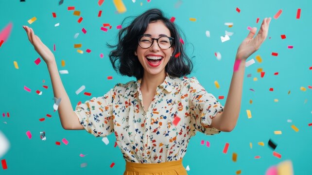 Young businesswoman with glasses and colorful shirt laughing with arms raised under a shower of confetti on a turquoise background manager accepts congratulations