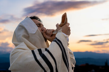 Young Jewish man blowing the shofar at sunset to celebrate the beginning of the Hebrew month or...