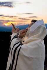 Young Jewish man blowing the shofar at sunset to celebrate the beginning of the Hebrew month or...