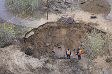 An aerial shot shows two workers in safety vests assessing a large sinkhole that has opened up near a path, disrupting the urban landscape. Concept of an urban infrastructure failure.