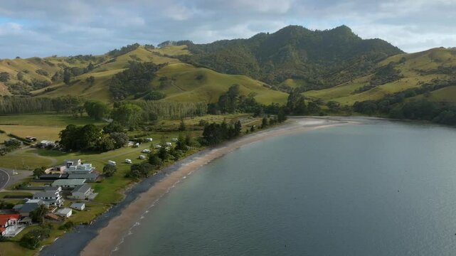 Aerial view of SIMPSON BEACH, WHITIANGA, COROMANDEL PENINSULA, New Zealand, nestled beside a bay. Houses and campervans line the beach. Rolling hills and lush greenery surround the tranquil scene.