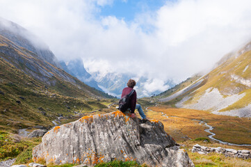 Person sitting on a rock in autumn, under clouds and a blue sky, seen from behind, looking at a...