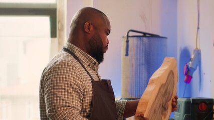 Fototapeta premium Portrait of jolly african american man inspecting wood before assembling furniture. Upbeat woodworking professional in studio evaluating lumber block, checking for damages, camera A
