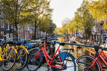 Fototapeta premium Colorful bicycles rest against the canal's edge as golden autumn leaves flutter in the breeze. The tranquil waterways of Amsterdam reflect the beautiful fall scenery, inviting exploration.
