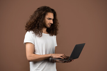 A young man with long curly hair stands alone, intently using a laptop. He is dressed casually in a white t-shirt, set against a warm brown background that enhances his concentration and lifestyle.