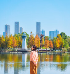 Fototapeta premium One Asian woman enjoying beautiful landscape of golden autumn season at Yinzhou Park, with lighthouse and urban CBD skyline in Ningbo, Zhejiang, China