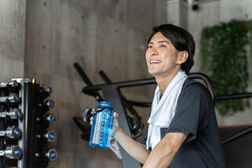 フィットネスジムで休憩・インターバル中に水・プロテインを飲む若いアジア人男性（水分補給）
