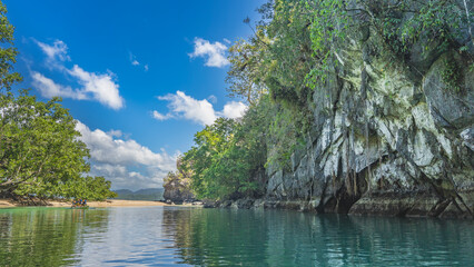 Sheer cliffs and a calm emerald river. There is green tropical vegetation on the sandy shore. Blue sky, clouds. Philippines. Palawan. Puerto Princesa Subterranean River National Park.