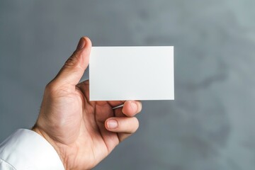 Obraz premium A Close-up of a Hand Holding a Blank White Business Card Against a Gray Background, Perfect for Mockups and Branding Presentations