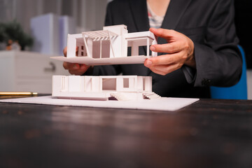 A salesman works late at night at his desk, surrounded by modern house models and drafts, analyzing...