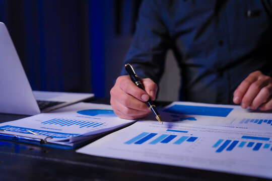 A man works late at night at his desk, hands close-up on charts and documents, conducting data analysis and strategic planning as a Policy and Planning Analyst focused on impactful decision-making. - Powered by Adobe
