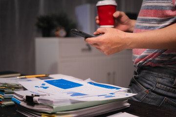 A man working late at home sits at his desk analyzing financial charts, checking stock market data on a laptop, tablet, and mobile, surrounded by informal, technology-driven tools.