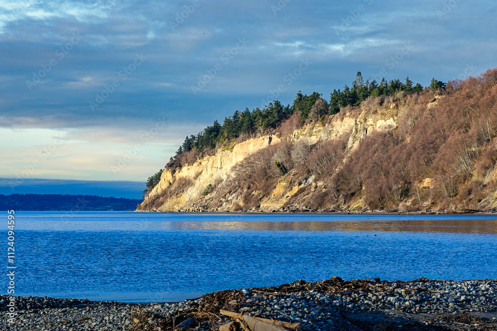 Wall mural 2023-12-31 PUGET SOUND WITH A ROCK COVERED SHORE IN THE FOREGROUND AND A NICE ROCKY CLIFF WITH TREES AND A NICE SKY ON WHIDBEY ISLAND WASHINGTON