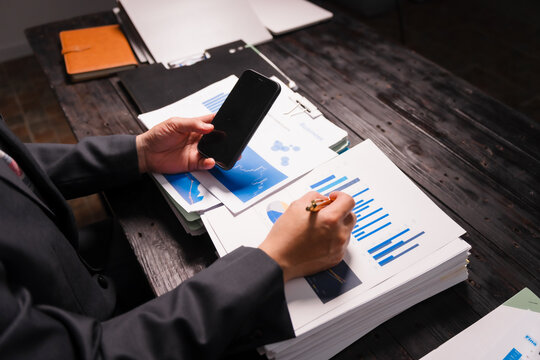 A young Asian man in a formal suit works late at night at his desk, analyzing stock market charts on a laptop and tablet, ensuring accuracy in financial data analysis.