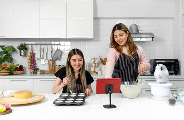 Two young women in an apron make dessert in the kitchen while looking at online recipe guide book on tablet computer.