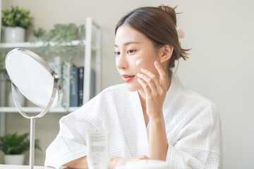 Facial beauty skin care, smile of pretty asian young woman in bathrobe looking at mirror, hand applying moisturizer lotion on her face, putting cream treatment before makeup cosmetic routine at home.