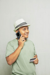 Adult Asian man wearing straw hat and light green shirt, answering phone call on his mobile phone, side view holding card, and smiling happily. Travel concept