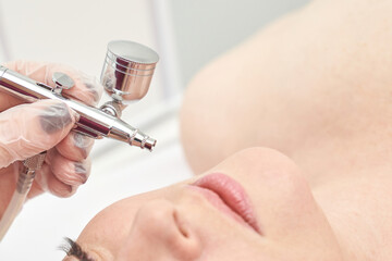 Caucasian female receiving professional facial treatment with airbrush device in spa setting.