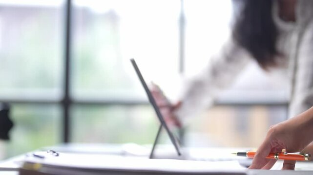 A person is pointing at a laptop screen with a pen. The scene suggests a collaborative work environment where people are working together on a project