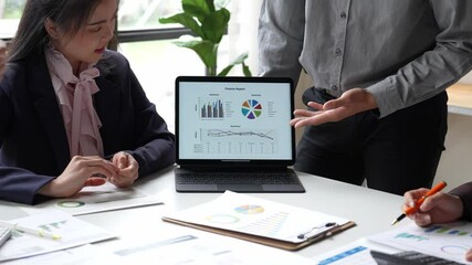 A group of people are looking at a laptop displaying graphs and charts. They are pointing at the screen and discussing the data. Scene is serious and focused