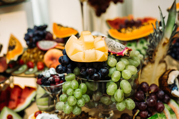 an assortment of various fruits, beautifully arranged on a glass plate. In the center is a coconut half filled with blueberries. Nearby lie pieces of mango, cut into cubes.
