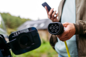 Close uf of man holding charger and smartphone in hands. Charging electric car in front of his...