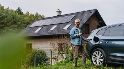 Man charging electric car, plugging the charger into the charging port. House with solar panel...