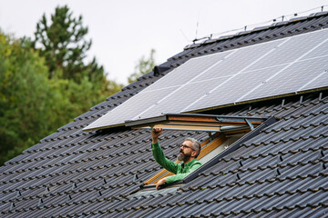 Mature man closing skylight, roof window, solar panels above him. Sustainable lifestyle and green...