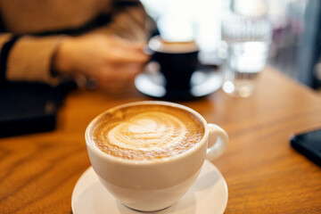 Close up of coffee cup with cappuccino with woman drinking her coffee in blurry background.