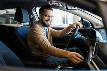 Young smiling customer sitting in new car in car showroom with hand on steering wheel and testing a car.