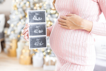 Young pregnant woman holding an ultrasound shot of her baby at home on Christmas Eve. Three photos fetus in the womb. Normal course of pregnancy. Third trimester December. successful ivf. surrogacy. 