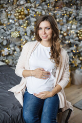 Young smiling white teeth beautiful Caucasian pregnant woman touching belly while celebrating Christmas at home looking at camera. The magic and magic of the winter holidays. Third trimester December