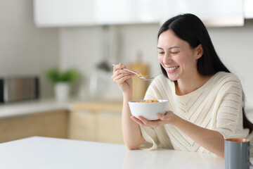 Happy asian woman eating cereals from bowl in the kitchen
