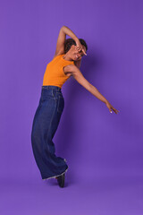 Young African American dancer in an orange top and denim skirt elegantly poses with a joyful expression. The vibrant purple background adds to the energy of this artistic moment captured in time.