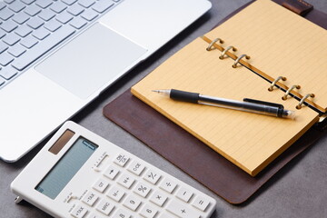 電卓とペン、システム手帳、ノートパソコンでデスクワークをしているイメージ
