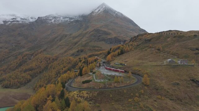 A picturesque alpine railway bends through the vibrant autumn landscape near Alp Gr&uuml;m, Graub&uuml;nden, Switzerland. The scene features a train, colorful foliage, and rugged mountain peaks