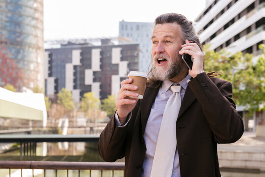 surprised mature male with beard in stylish elegant suit with cup of takeaway coffee in hand talking on mobile phone while standing on urban street, concept of business and mature man lifestyle