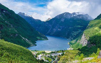 A scenic view of a Norwegian fjord surrounded by towering green mountains. The water is calm and blue, and the sky is clear. Geiranger fjord Norway in summer