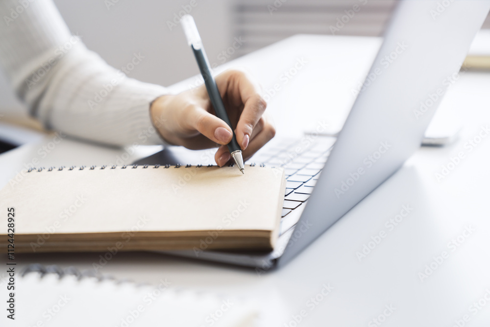 Poster the image focuses on a woman's hand scribbling in a notepad on a stylish laptop, set against a blurr