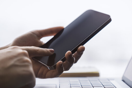 Woman engaged with her modern smartphone, laptop slightly out of focus nearby against the blurred office hustle and bustle