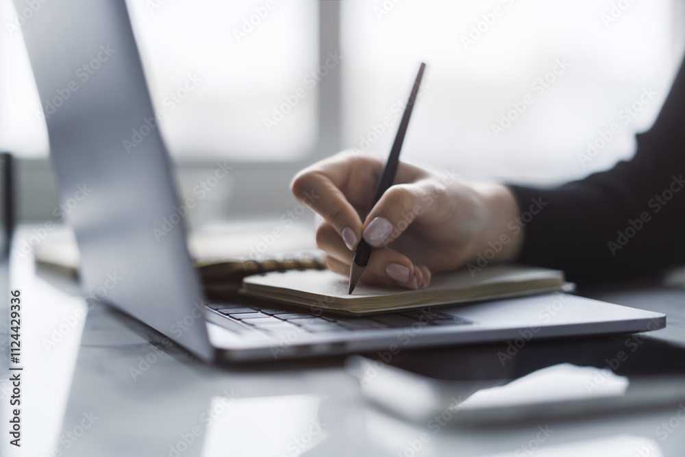 Poster the close-up image showcases a woman's hand inscribing in a notepad on a modern laptop, with a soft-
