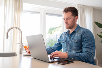 Fototapeta premium Working at home in the kitchen. caucasian male sitting at the kitchen counter behind his laptop working and communicating. Wearing a denim shirt with serious face expression.