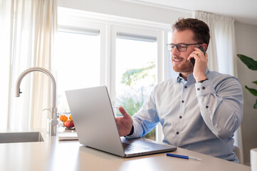 Working at home in the kitchen. Caucasian male sitting at the kitchen counter behind his laptop working and communicating. Wearing a light blue formal shirt and glasses