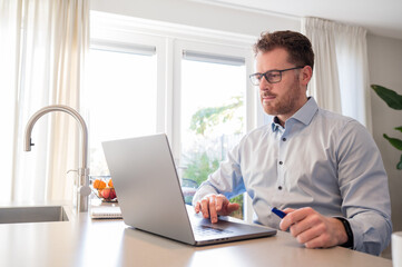 Working at home in the kitchen. Caucasian male sitting at the kitchen counter behind his laptop working and communicating. Wearing a light blue formal shirt and glasses.