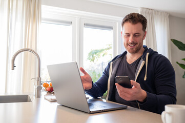 working at home in the kitchen. caucasian male sitting at the kitchen counter behind his laptop working and communicating. Dressed in a hoodie, holding phone in the hand on a video call.