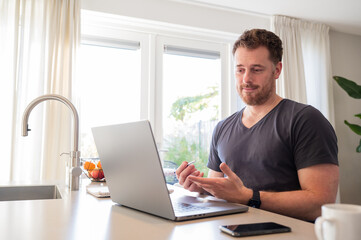 working at home in the kitchen. caucasian male sitting at the kitchen counter behind his laptop working and communicating. Wearing simple grey t shirt. On a conference call, making gestures with hands