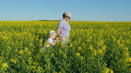 Fototapeta premium Mom Walking With Baby In Rapeseed Field. Girl With A Child Walking Along A Blooming Rapeseed. Together In Nature. Mother Day Concept.