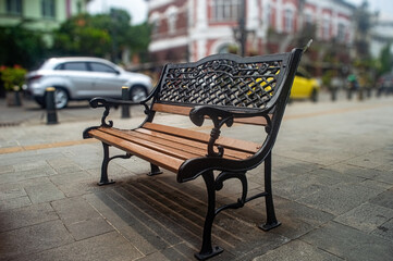 A beautifully designed park bench with intricate wrought iron armrests and backrest, set on a paved street with blurred background of cars and buildings, creating a serene urban scene